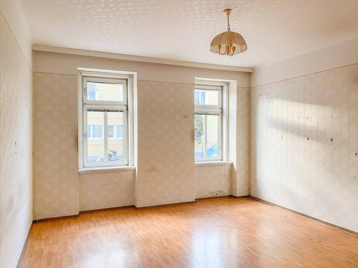 Heller Raum mit zwei Fenstern, Parkettboden und alter Tapete, bereit zur Renovierung.