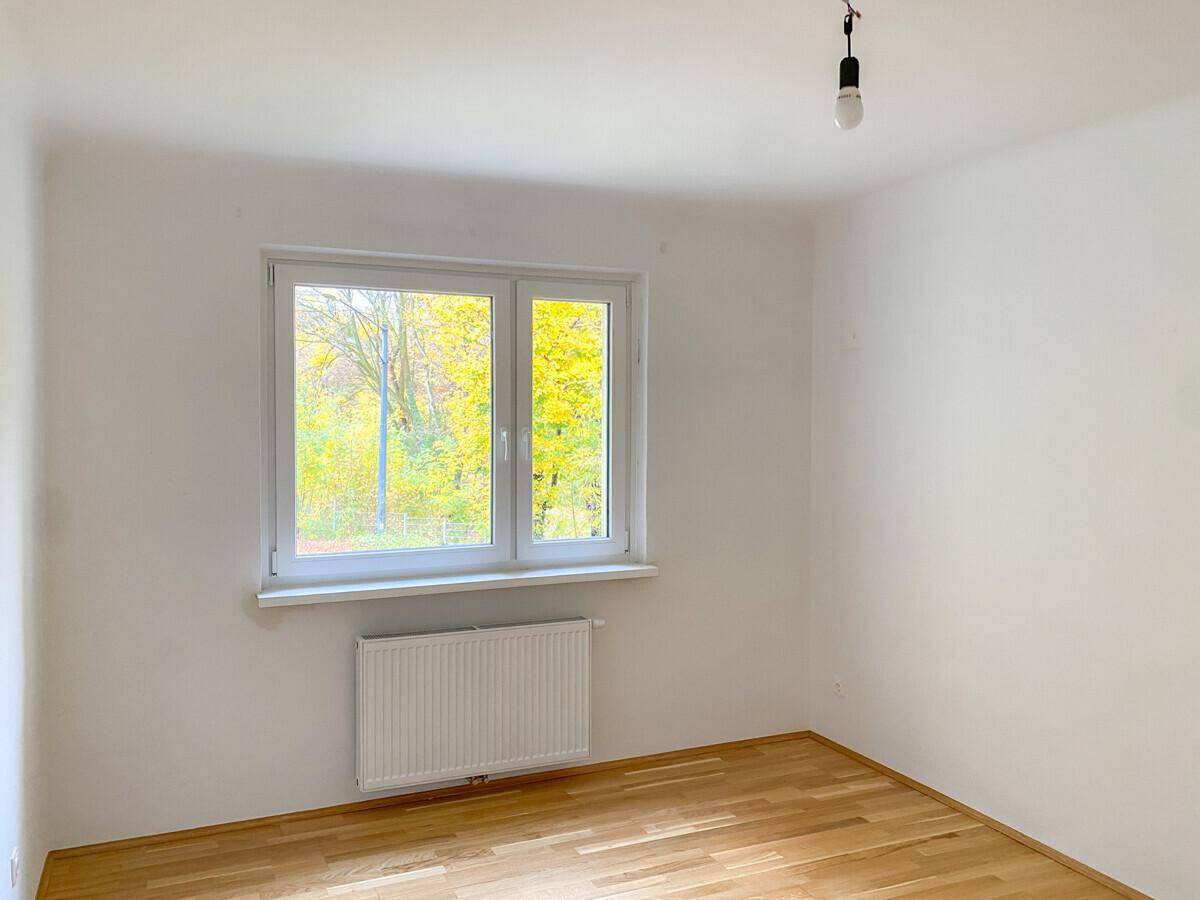 Lichtdurchflutetes Zimmer mit hellem Parkettboden, weißen Wänden und großem Fenster mit Blick ins Grüne.