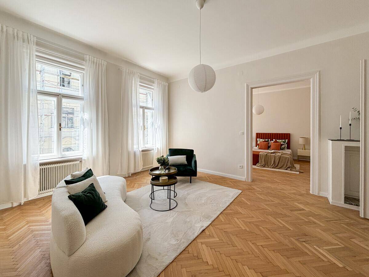 Geräumiges Wohnzimmer mit elegantem Fischgrätparkett und Blick in ein angrenzendes Schlafzimmer.