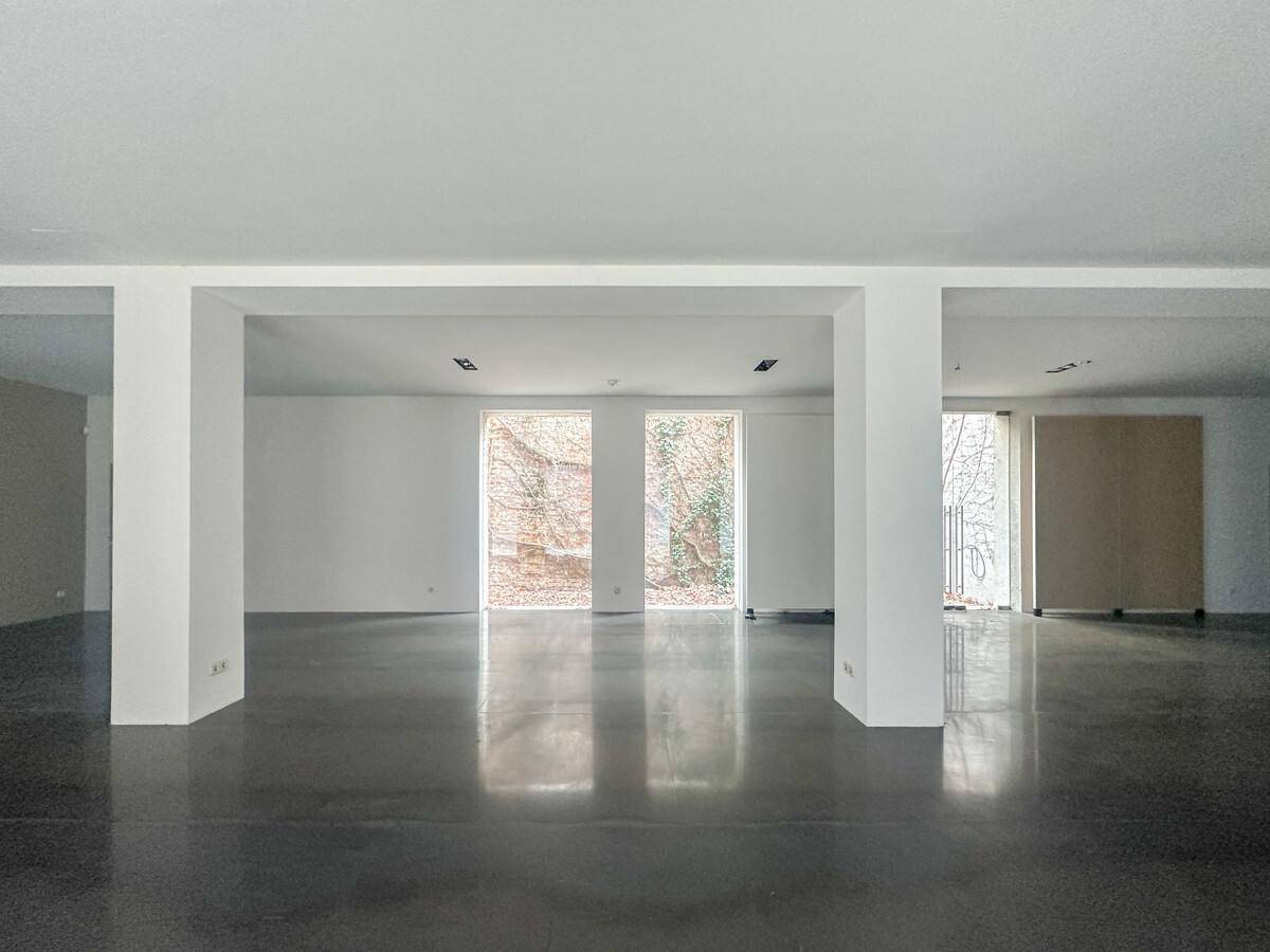 Geräumiger, heller Innenraum mit Betonboden und großen Fenstern, die viel Tageslicht und Grünblick bieten.