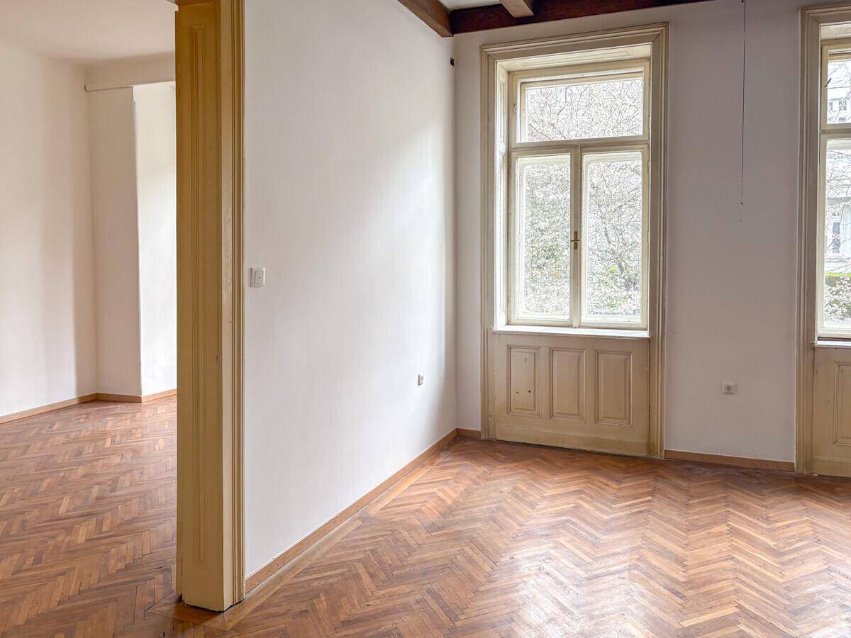 Helles Zimmer mit Fischgrätparkett, hohen Fenstern und sichtbaren Deckenbalken.