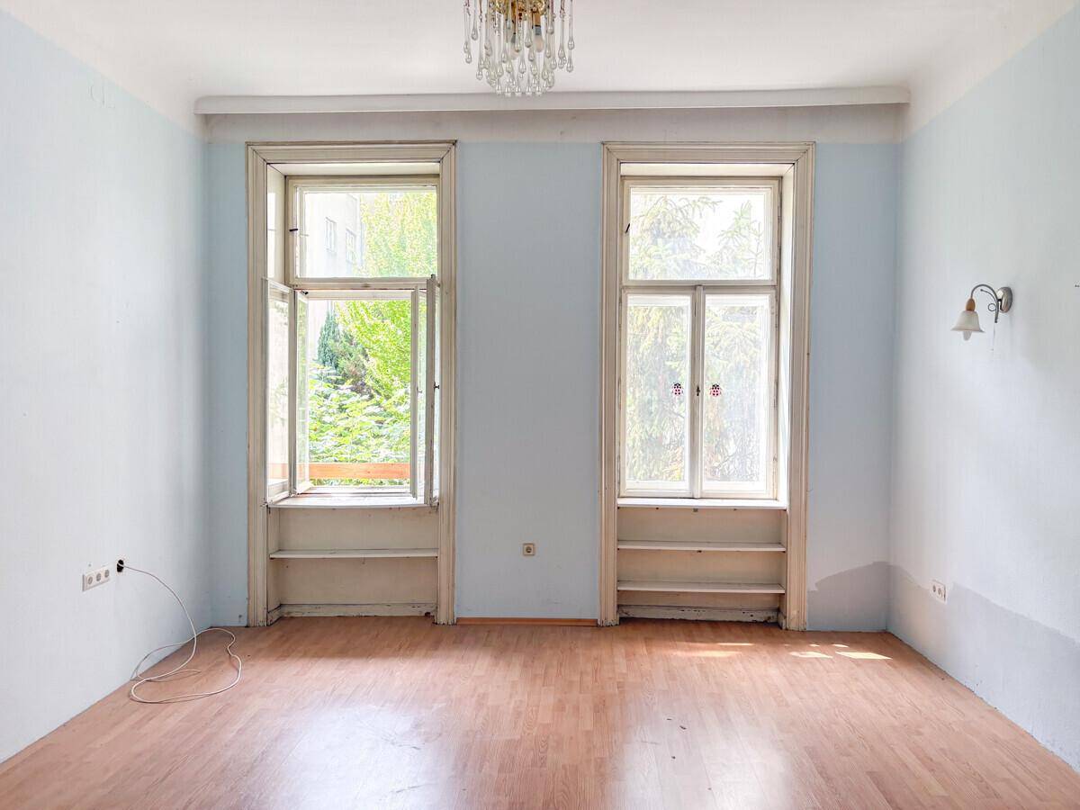 Leerer Raum mit hellblauen Wänden, Laminatboden und zwei großen Fenstern mit Blick ins Grüne.