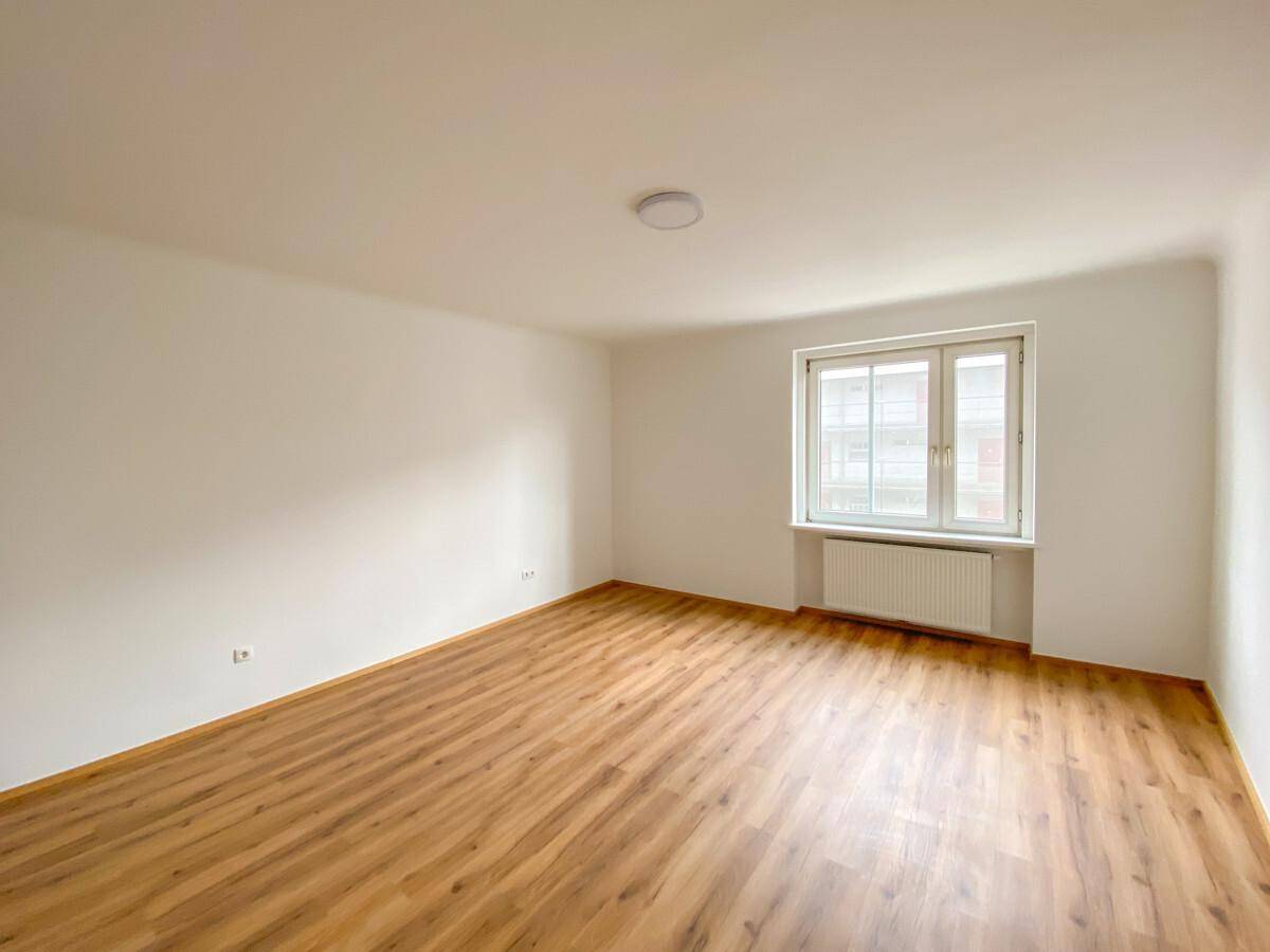 Lichtdurchflutetes, leeres Zimmer mit Fenster, Heizkörper und warmem Laminatboden.