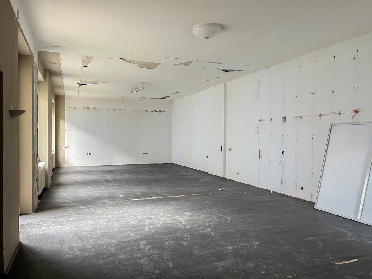 Großer, leerer Raum in Renovierung mit abblätternder Decke und dunklem Bodenbelag.