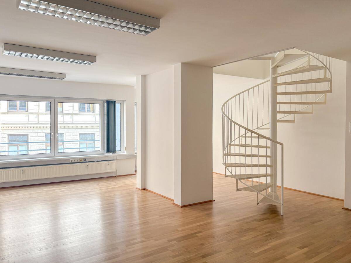 Heller Büroraum mit Holzparkettboden, großen Fenstern und einer eleganten Wendeltreppe, die in eine obere Etage führt.