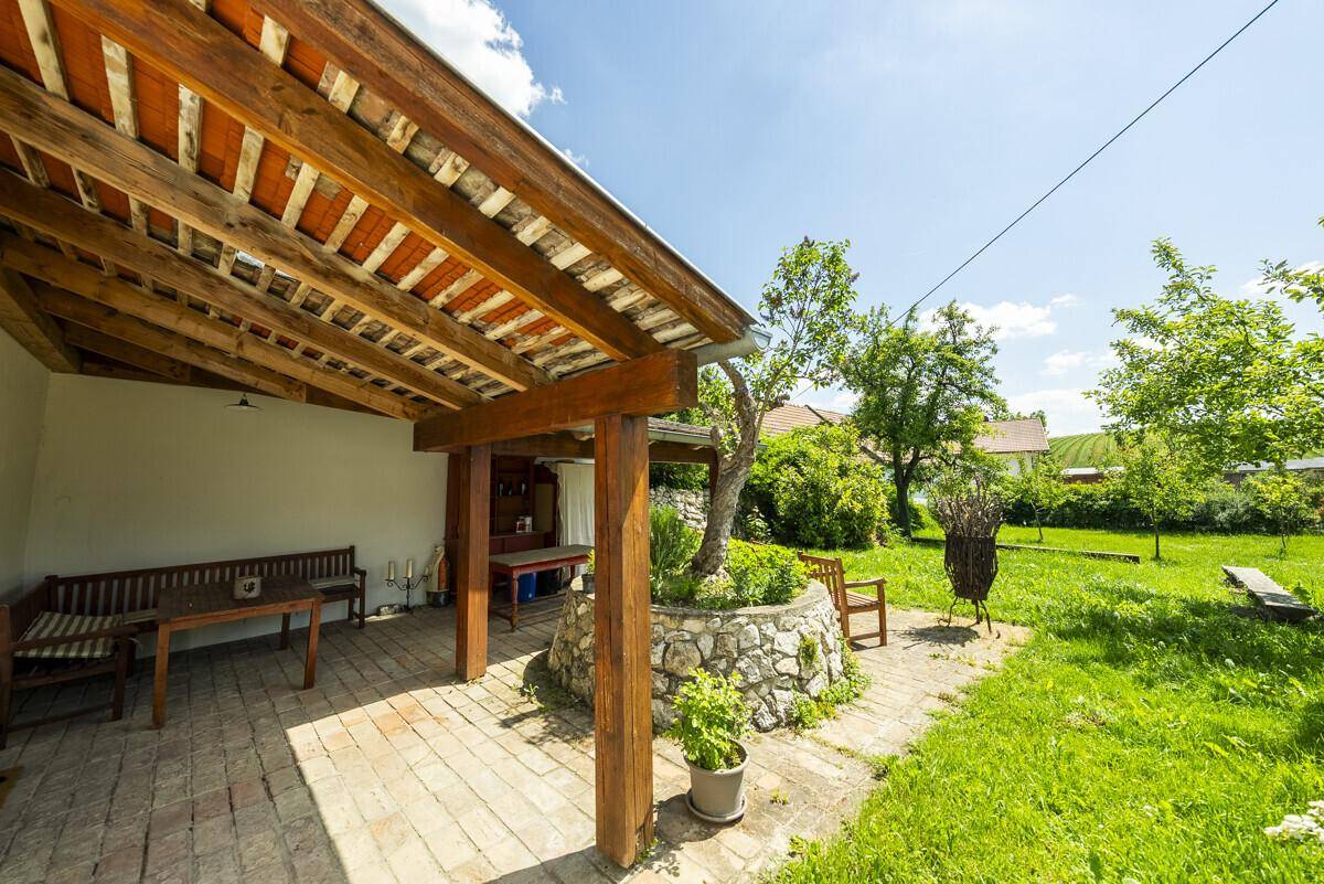 Überdachter Sitzbereich im Freien mit Holzbalken, gepflastertem Boden und Blick auf den Garten.