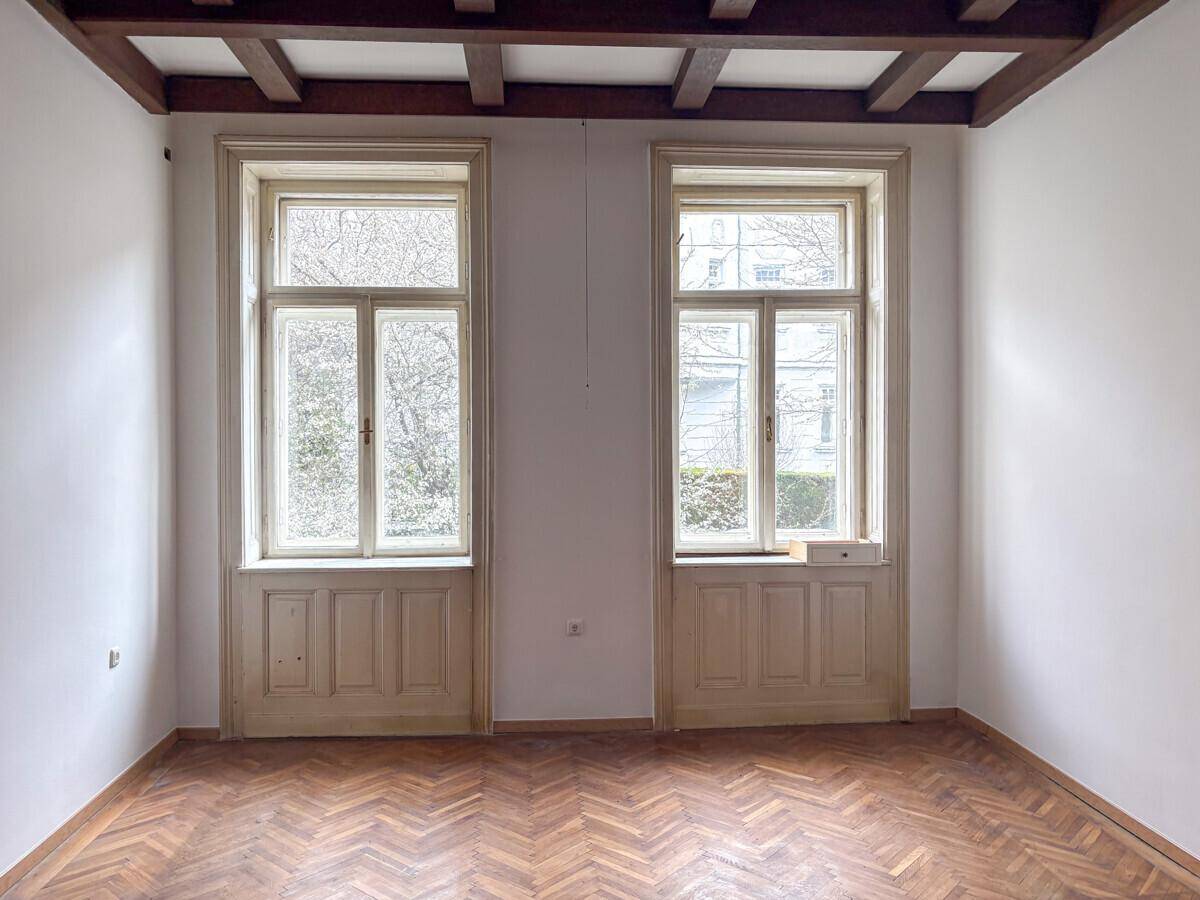Zimmer mit zwei hohen Fenstern, Fischgrätparkett und sichtbaren Holzbalken an der Decke.