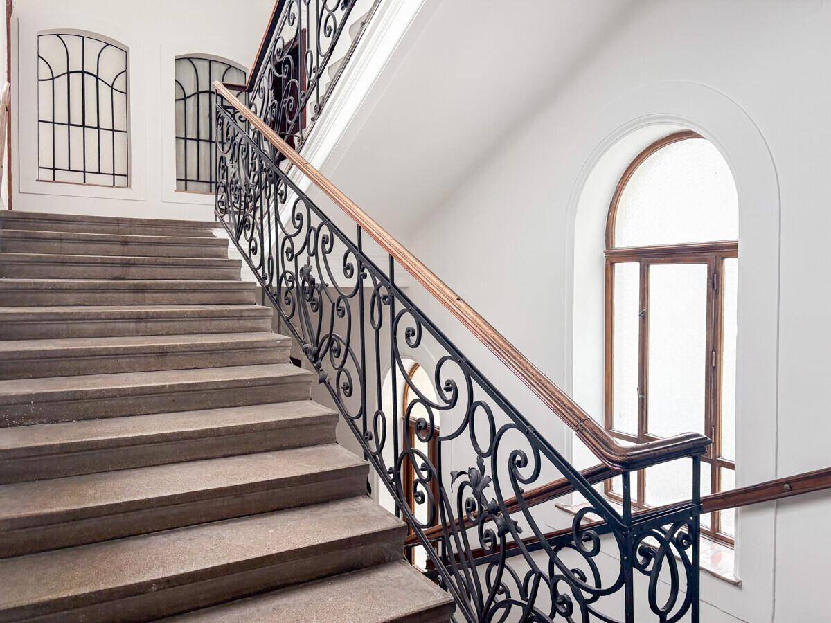 Historisches Treppenhaus mit Steinstufen, schmiedeeisernem Geländer und großen Bogenfenstern.