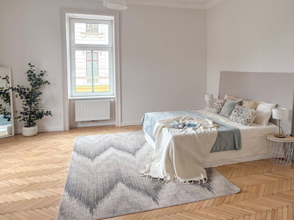 Helles Schlafzimmer mit großem Bett, Fischgrätparkett und einem großen Fenster.