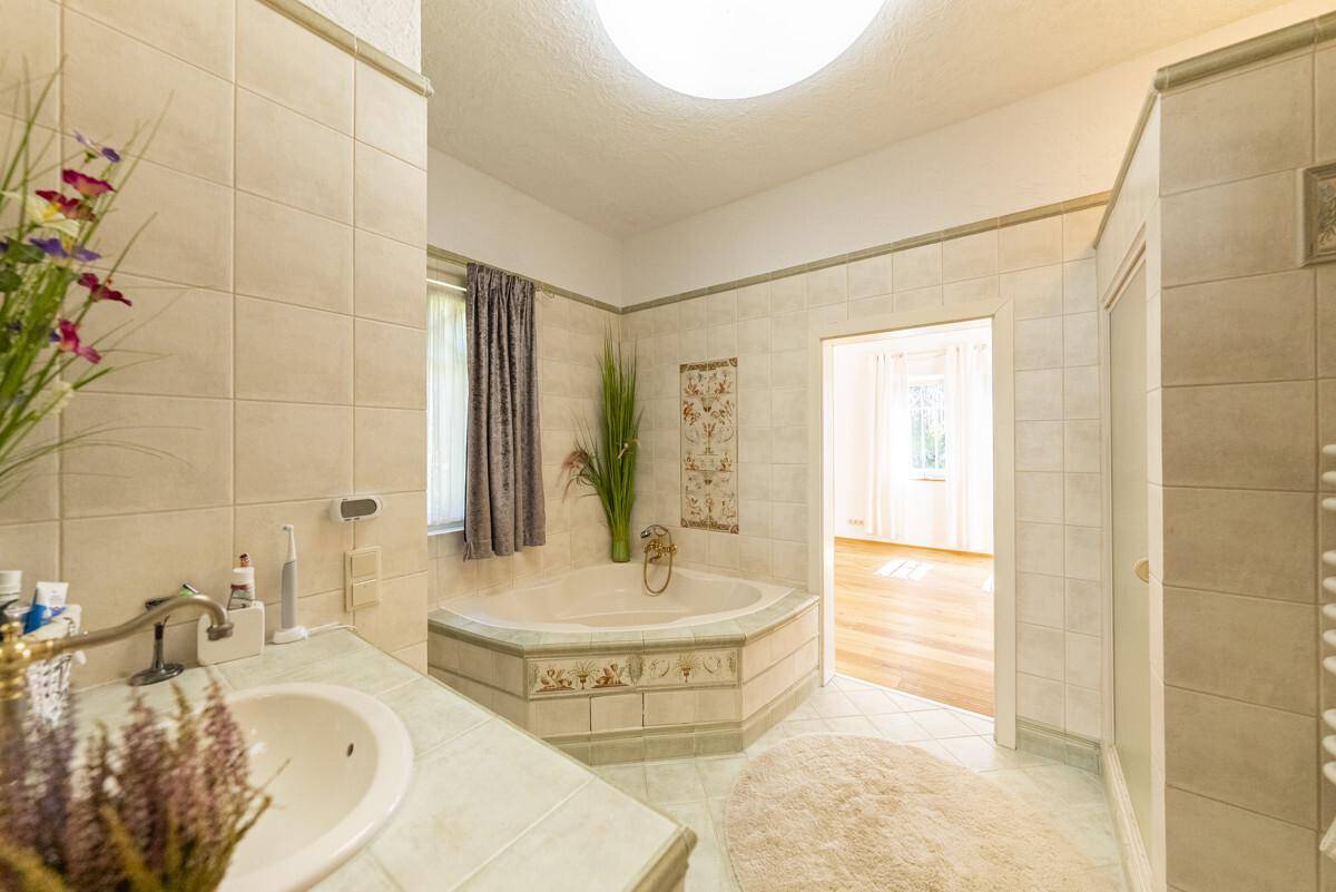 Helles Badezimmer mit Eckbadewanne, beigefarbenen Fliesen und einem Fenster für Tageslicht.