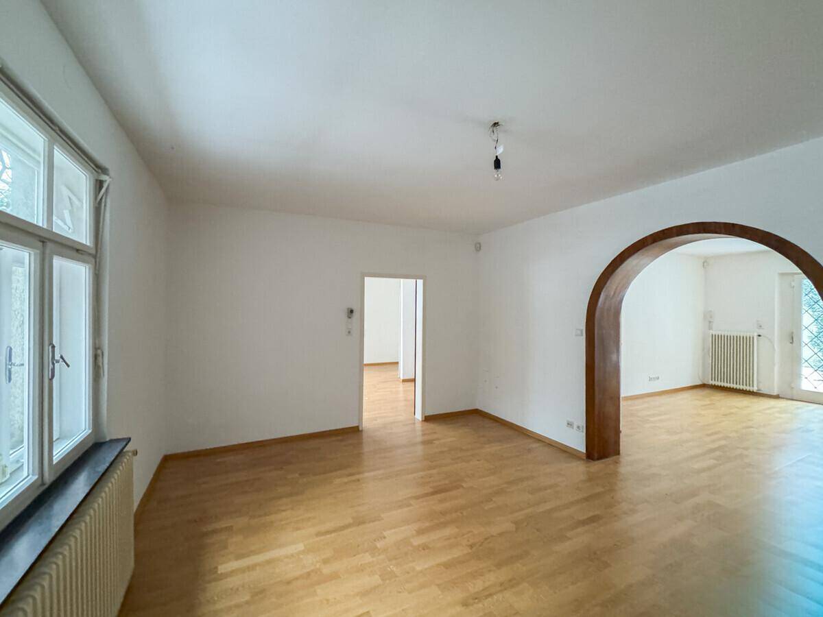 Geräumiges Wohnzimmer mit Parkettboden, großen Fenstern und einem Rundbogen zu einem angrenzenden Bereich.