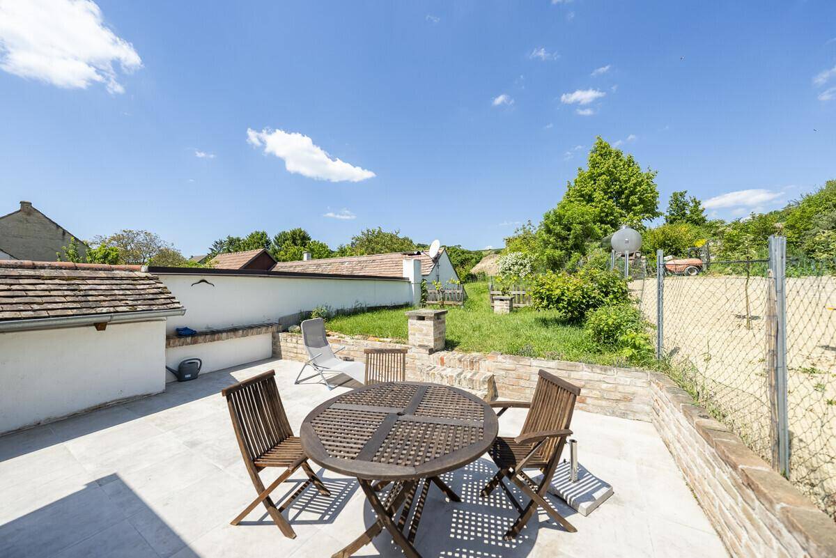 Sonniger Außenbereich mit gepflasterter Terrasse, Holztisch und Stühlen, umgeben von Grünflächen.