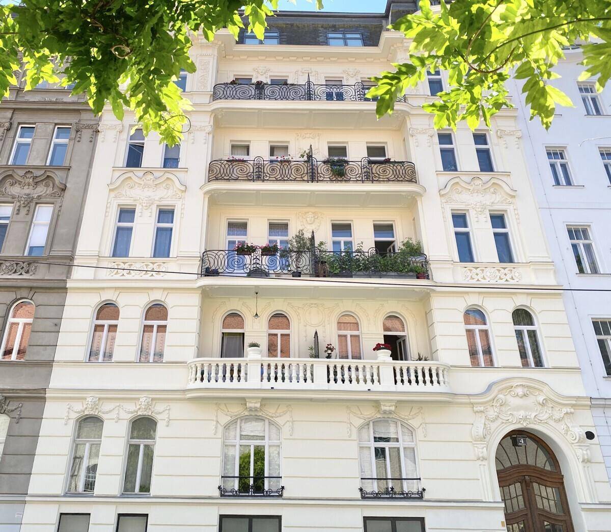 Elegante Altbaufassade mit reich verzierten Balkonen und großen Fenstern, umgeben von grünen Blättern.