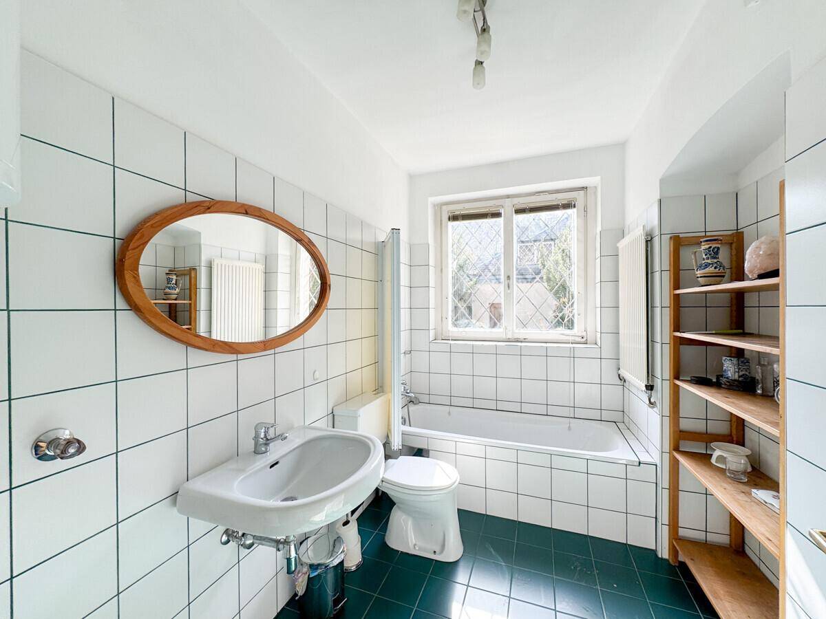Badezimmer mit weißen Fliesen, Badewanne, Fenster und einem ovalen Holzspiegel an der Wand.