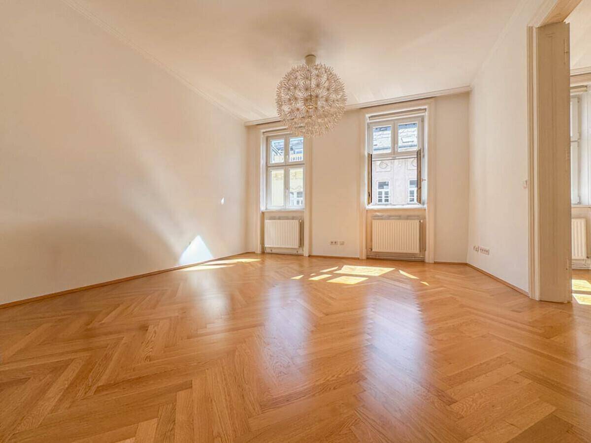 Geräumiges Wohnzimmer mit Fischgrätparkett, hohen Decken und großen Fenstern, die viel Licht hereinlassen.