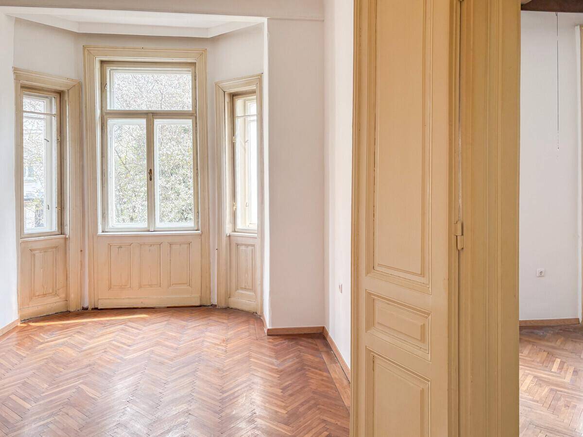 Lichtdurchfluteter Raum mit Erkerfenstern, Fischgrätparkett und einer traditionellen Holztür.