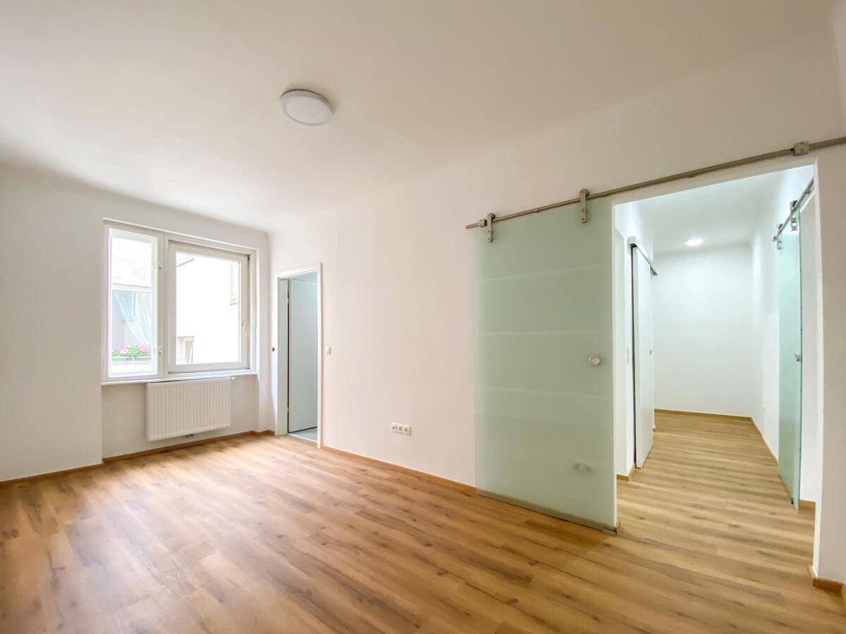 Helles, leeres Zimmer mit Laminatboden, weißen Wänden und moderner Glasschiebetür.