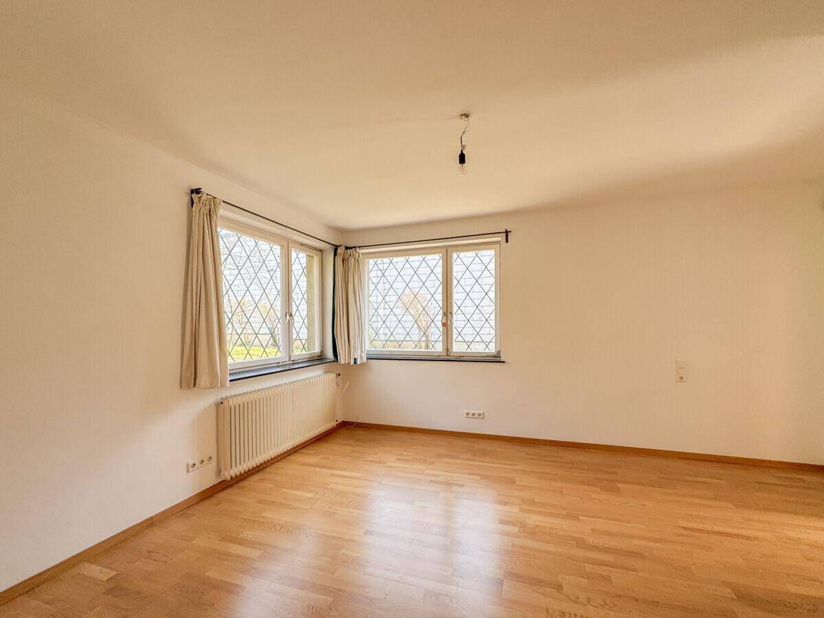 Helles, leeres Zimmer mit Parkettboden und großen Fenstern, die viel Tageslicht hereinlassen.