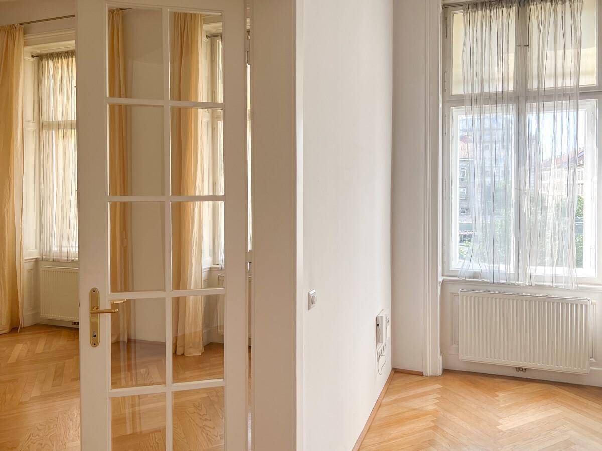 Helles Zimmer mit Fischgrätparkett, hohen Fenstern und Vorhängen.