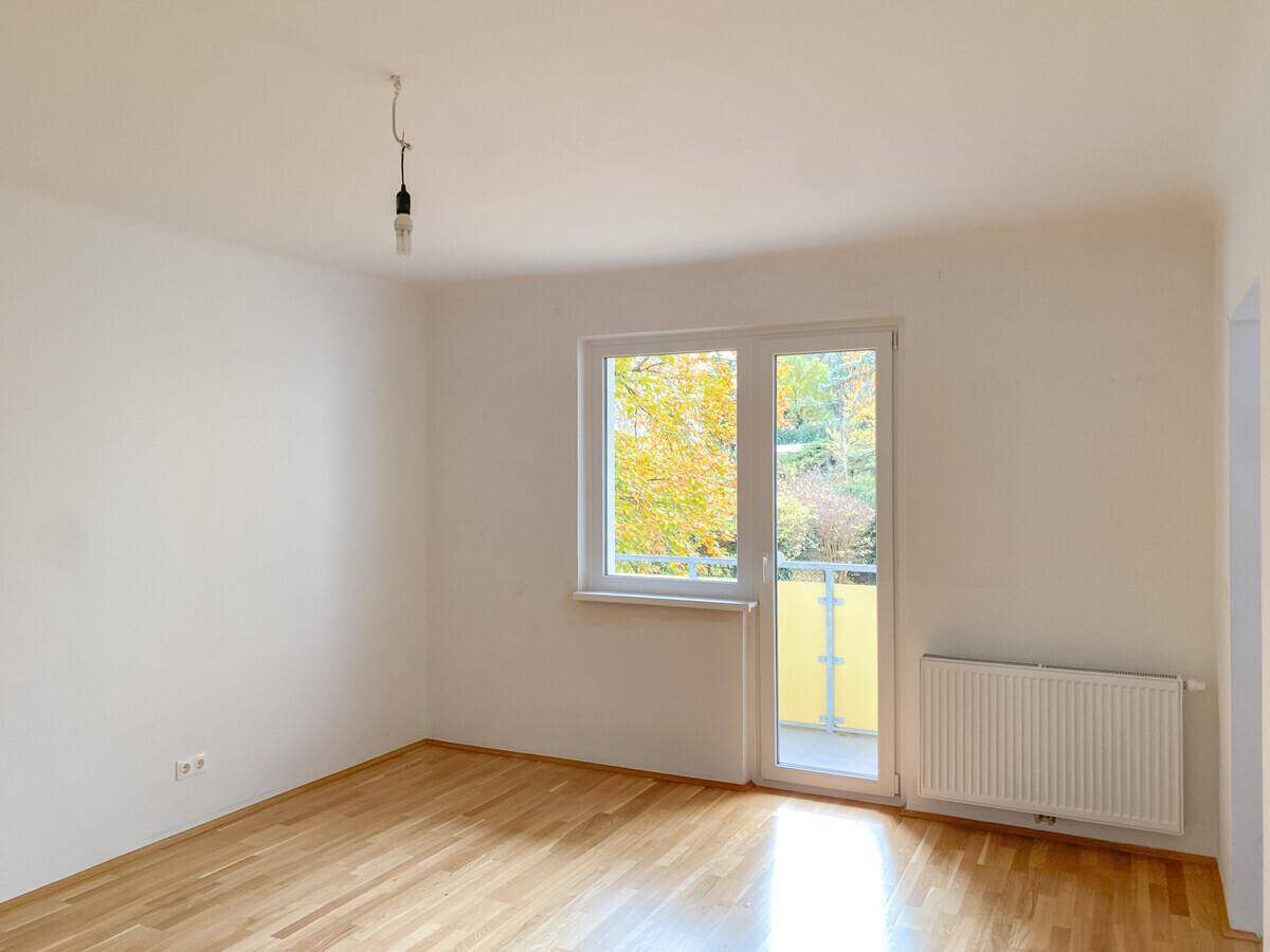 Heller Raum mit Parkettboden, weißen Wänden und Zugang zu einem Balkon mit Blick ins Grüne.