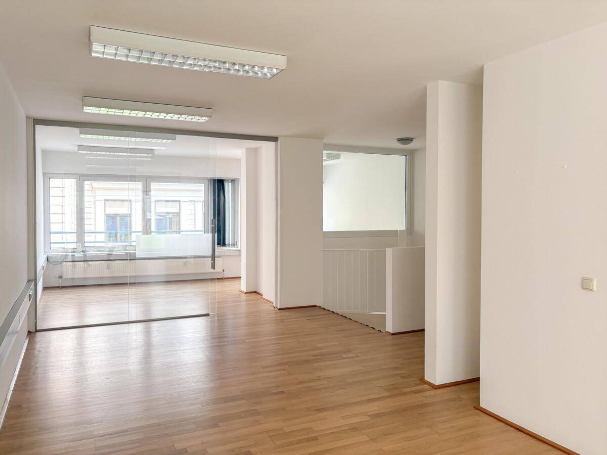 Offener Bürobereich mit Holzparkettboden und einer Glaswand, die einen weiteren hellen Raum abtrennt.