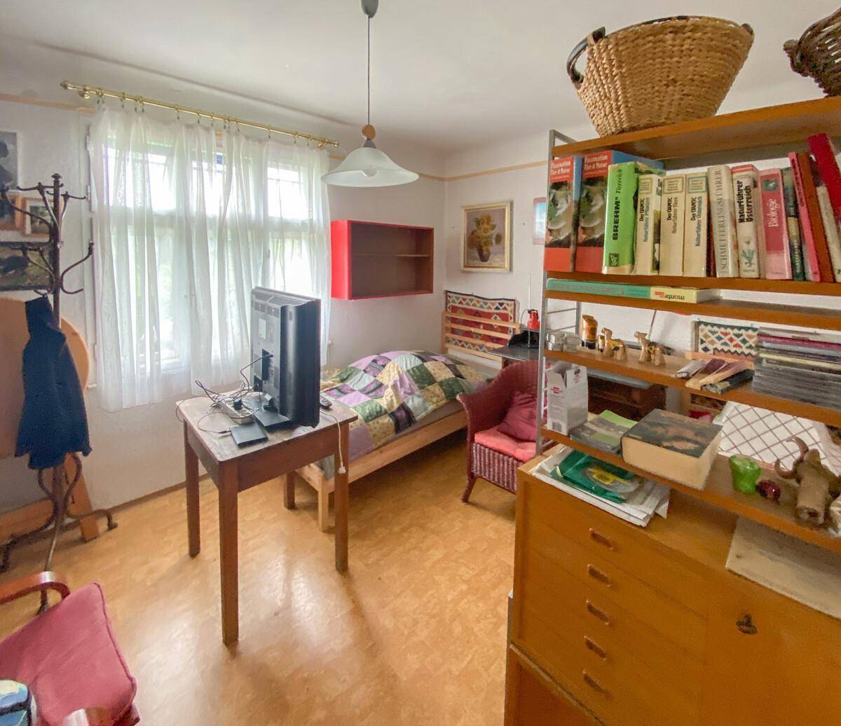Schlafzimmer mit Einzelbett, Fenster und einem Bücherregal, einfach eingerichtet.