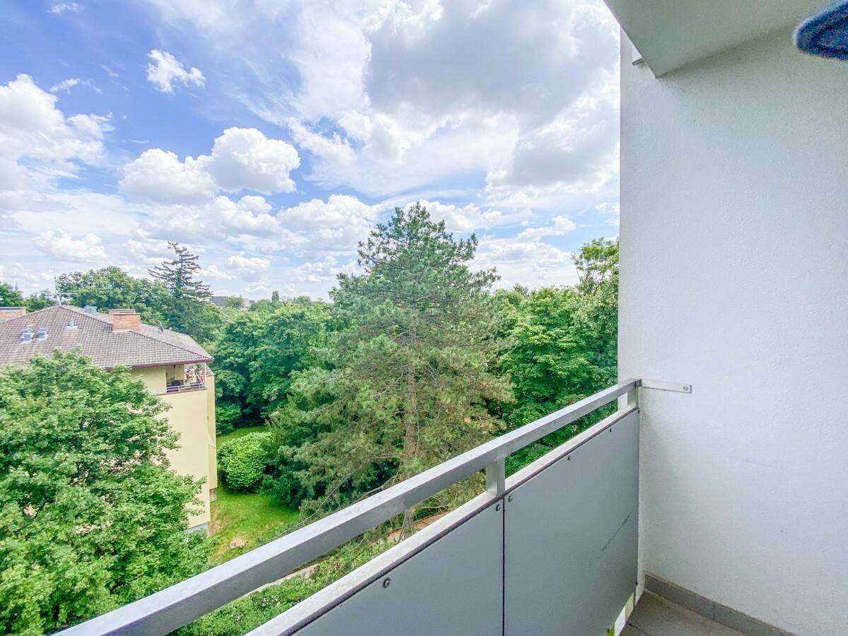 Balkon mit Ausblick auf grüne Bäume und Dächer von Wohnhäusern unter einem hellen, bewölkten Himmel.