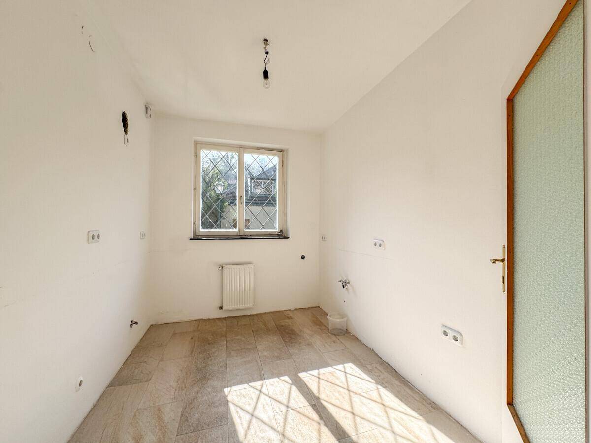 Leerer Küchenraum mit Fliesenboden, Fenster und Anschlüssen für eine zukünftige Einbauküche.