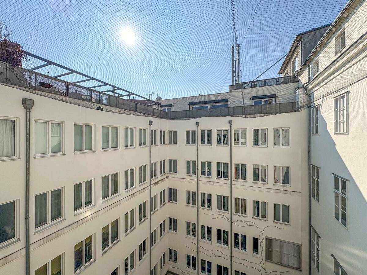 Blick in einen hellen Innenhof eines Gebäudes mit vielen Fenstern und einem Schutznetz darüber.