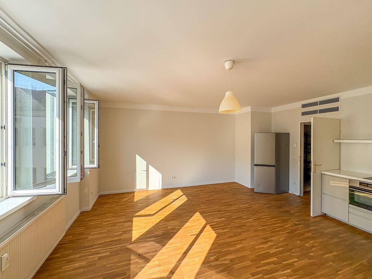 Helles Wohnzimmer mit Parkettboden, offener Küchenzeile und viel natürlichem Licht durch die geöffneten Fenster.