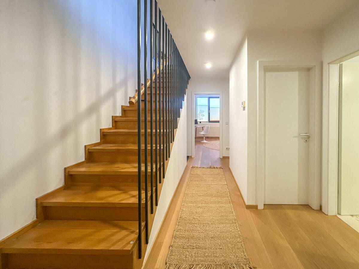 Heller Flur mit Holztreppe und modernem Geländer, der zu weiteren Räumen führt.
