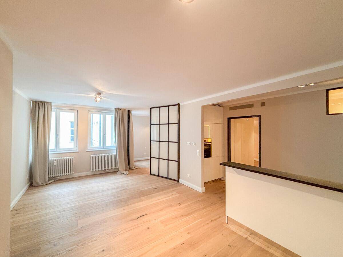 Geräumiger Wohnbereich mit großen Fenstern, hellem Eichenparkett und einer modernen Raumtrennung aus Glas.
