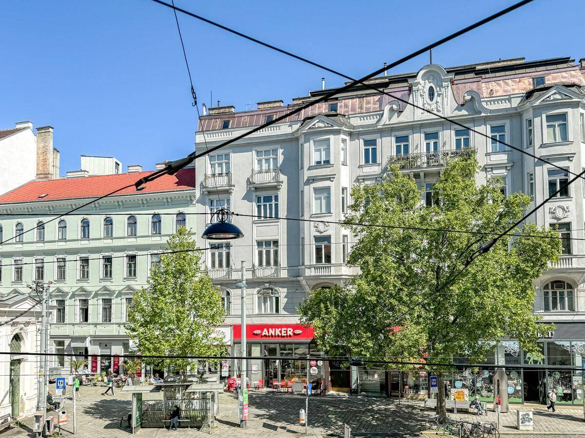 Historische Stadtfassade mit Balkonen, Geschäften im Erdgeschoss und grünen Bäumen an der Straße.