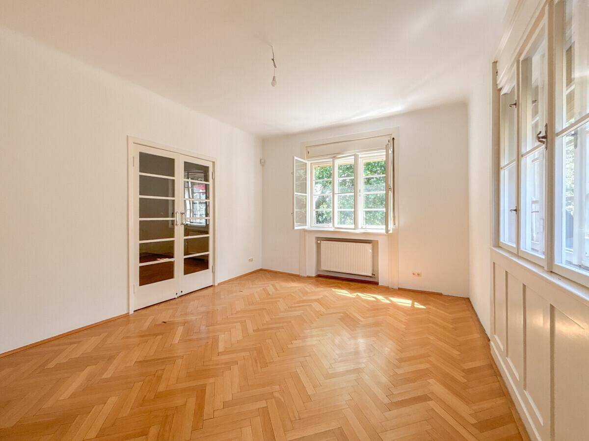 Geräumiges Zimmer mit Fischgrätparkett und großen Fenstern mit Blick ins Grüne.