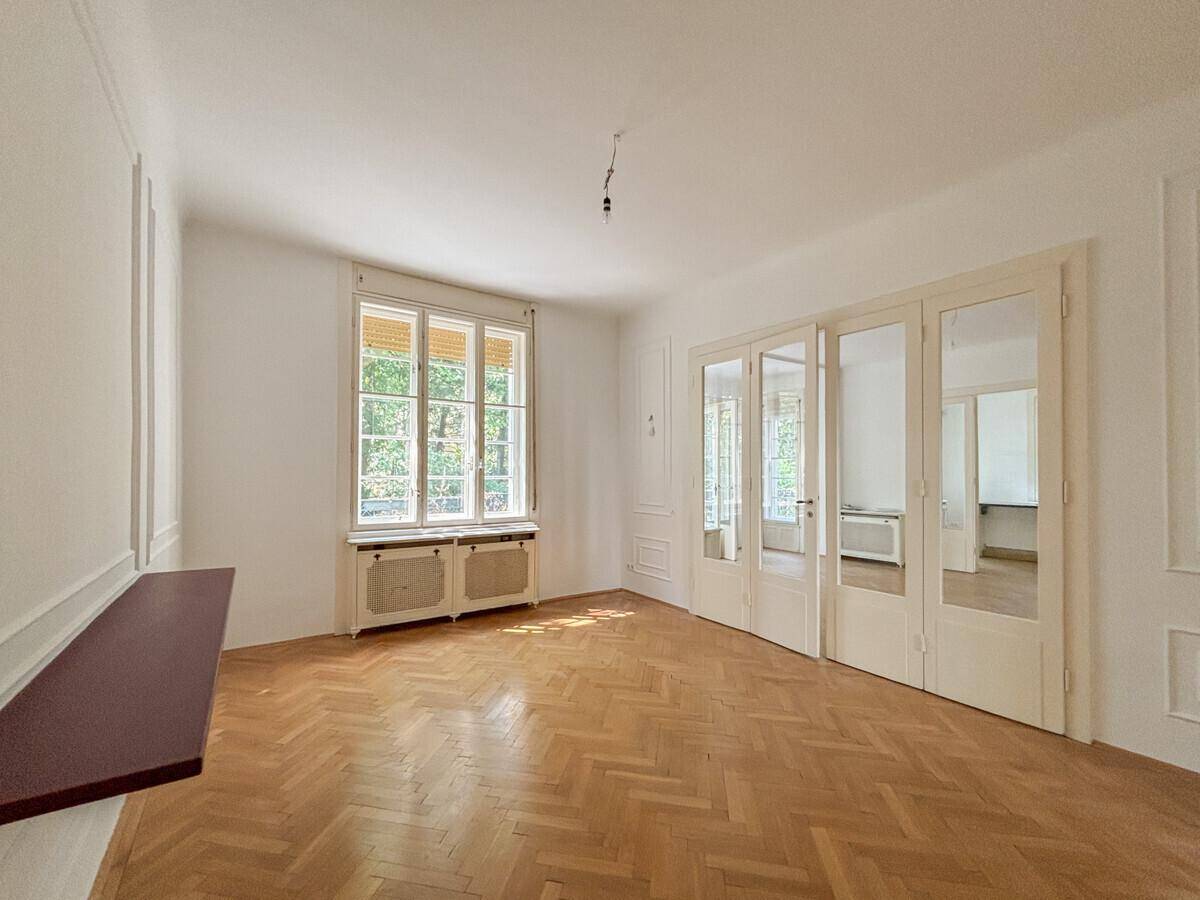 Helles Zimmer mit Fischgrätparkett, hohen Fenstern und klassischer Wandverkleidung.