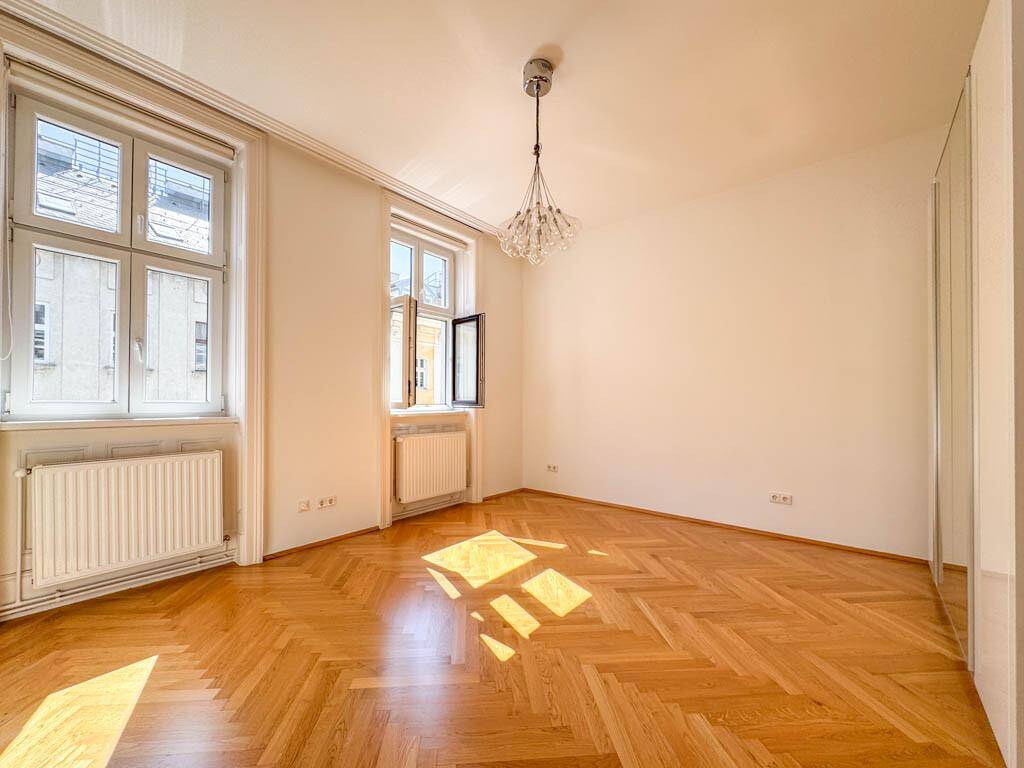 Lichtdurchflutetes Zimmer mit Fischgrätparkett, hohen Decken und einem modernen Kronleuchter.