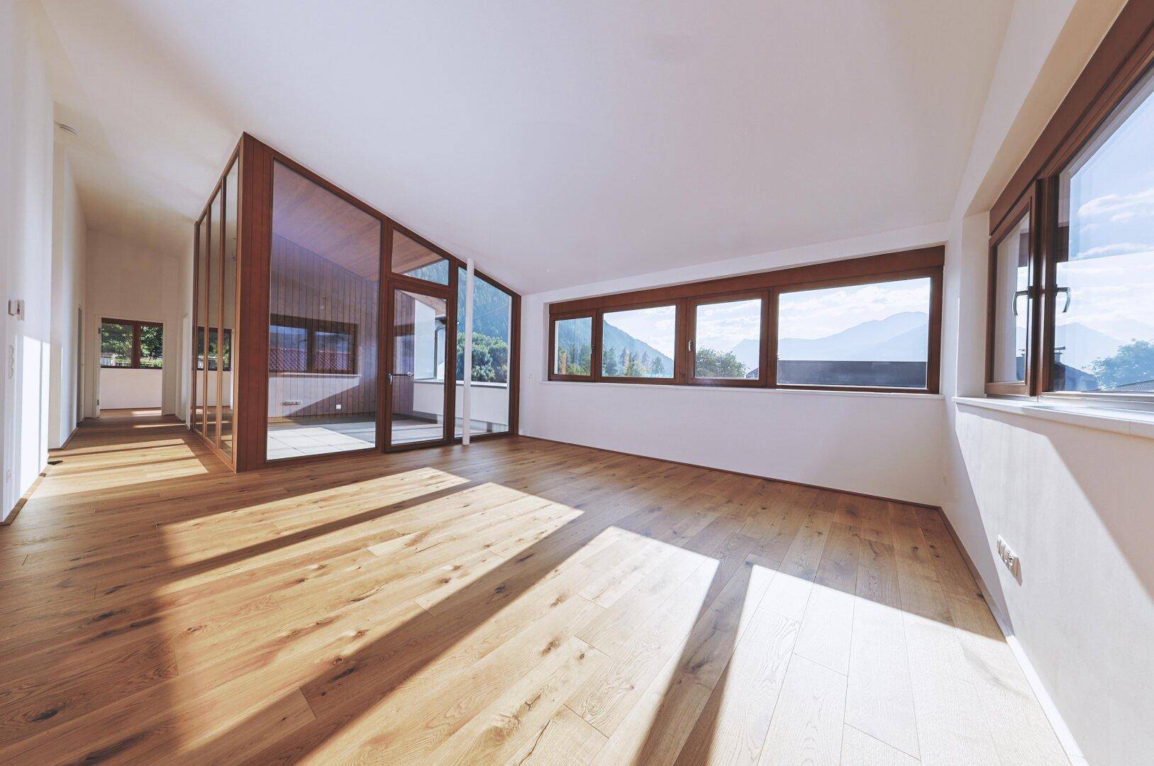 Geräumiger, leerer Wohnbereich mit Holzboden und großen Fenstern, die viel Tageslicht hereinlassen.