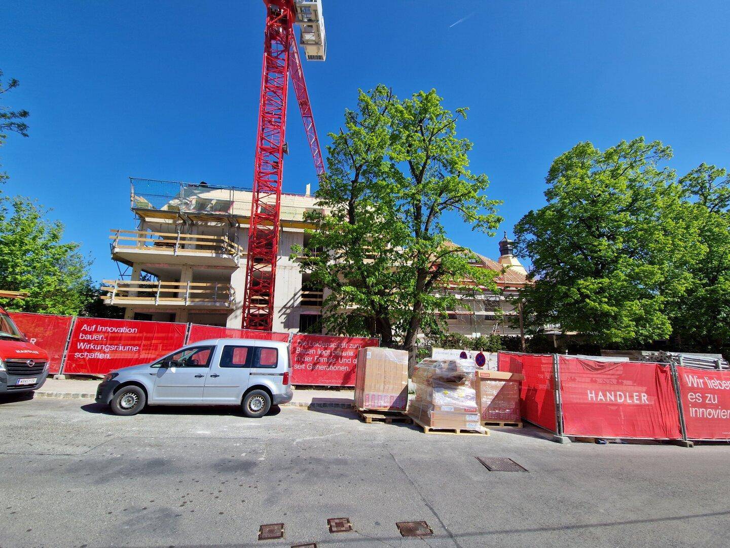 Baustelle mit einem im Bau befindlichen Gebäude, einem großen roten Kran und umgebenden Bäumen unter blauem Himmel.