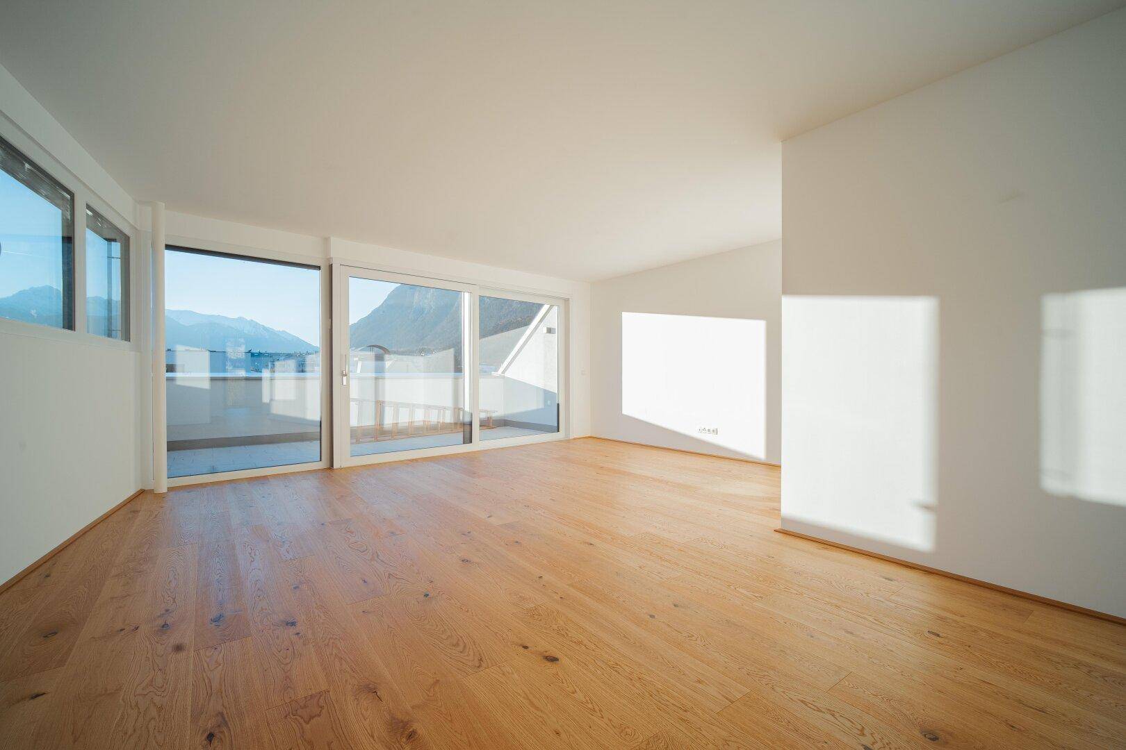 Geräumiges, leeres Wohnzimmer mit hellem Holzfußboden und großen Schiebetüren zur Terrasse mit Bergblick.