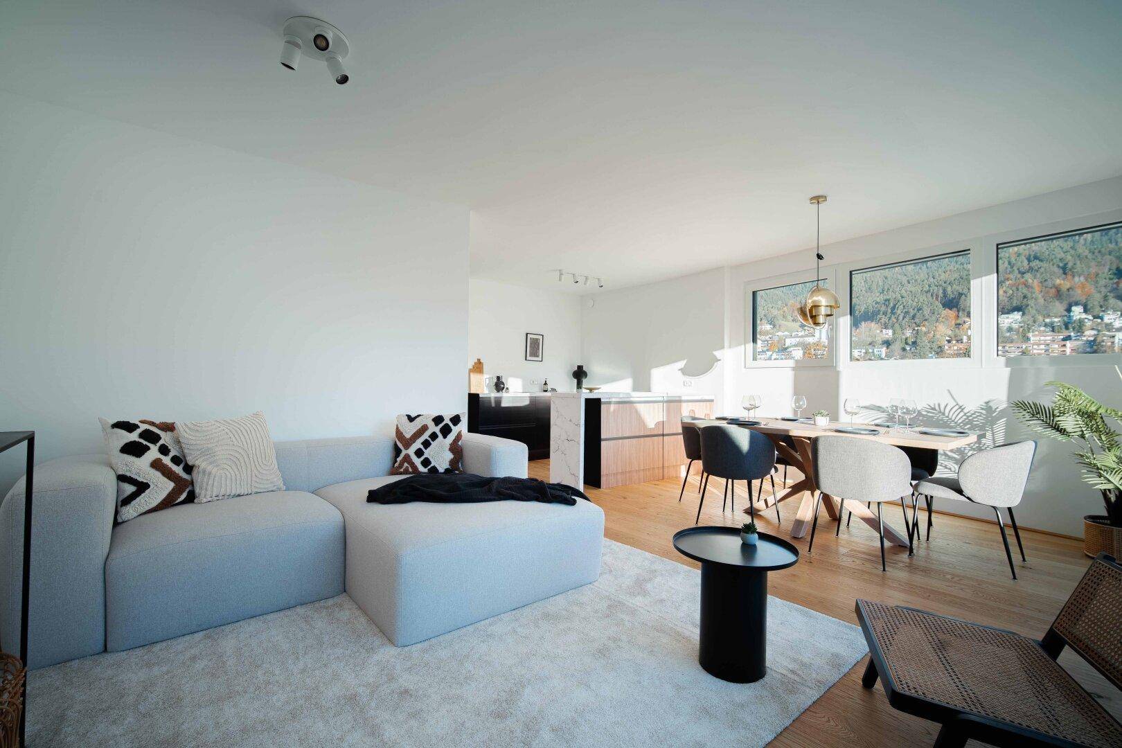 Moderner offener Wohn- und Essbereich mit bequemer Couch, Esstisch und integrierter Küche im Hintergrund.