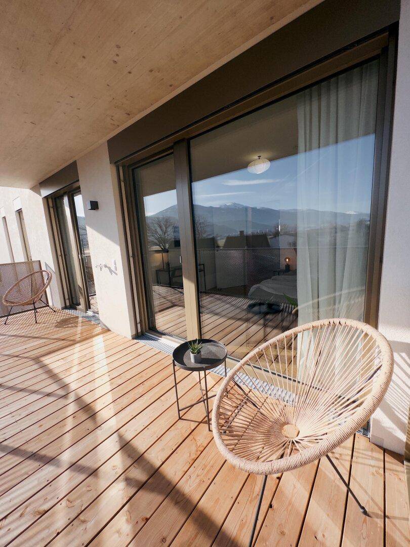 Sonniger Balkon mit Holzboden und gemütlichen Sitzgelegenheiten, ideal zum Entspannen mit Aussicht.