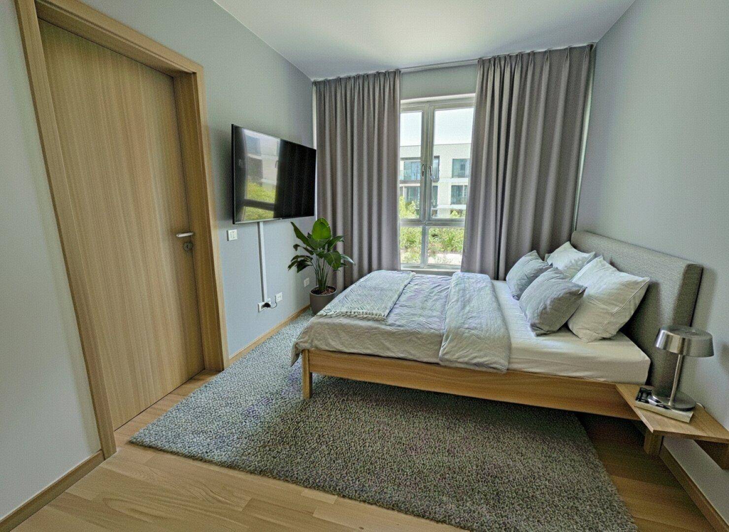 Helles Schlafzimmer mit Holzboden, großem Fenster, Wandfernseher und modernen Vorhängen.
