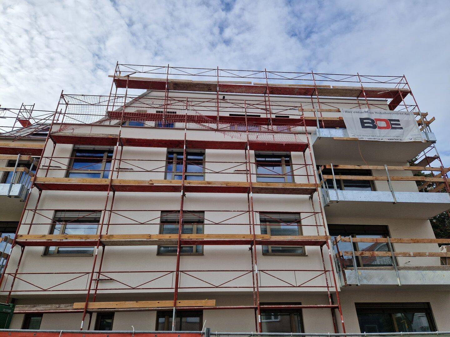 Nahaufnahme der modernen Fassade eines Neubaus mit Gerüst, großen Fenstern und Balkonen.