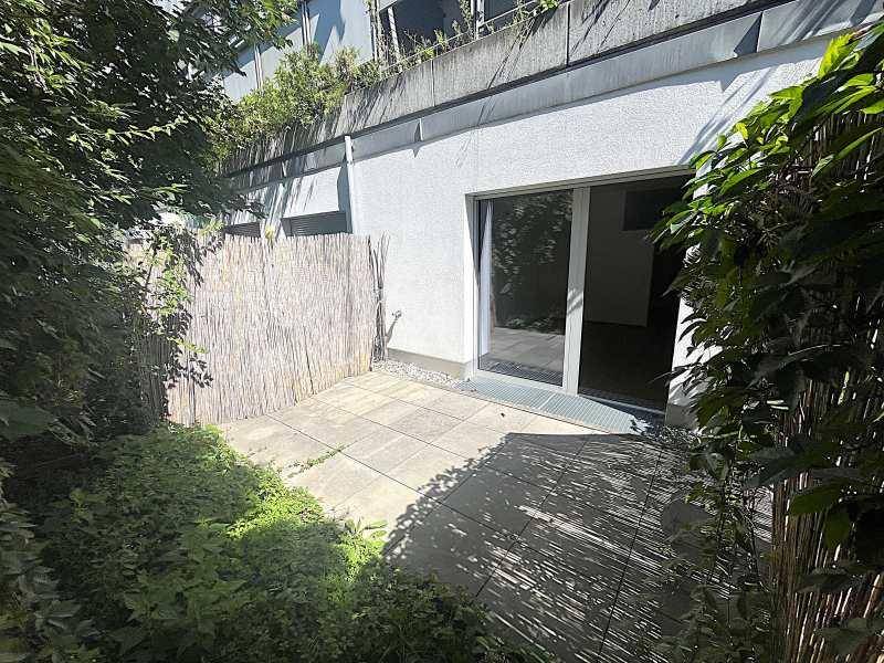 Kleine gepflasterte Terrasse im Freien, umgeben von Grünpflanzen und einer Sichtschutzwand.