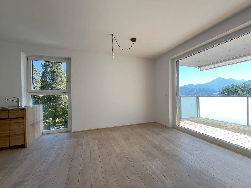 Geräumiger, leerer Wohnbereich mit Holzboden, großen Fenstern und Bergblick vom Balkon.