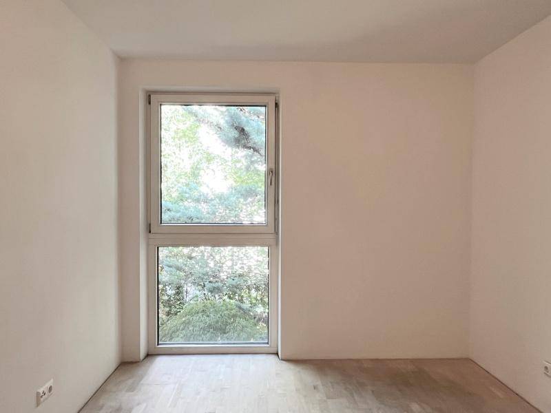 Leeres Zimmer mit Holzboden und einem großen Fenster, das einen grünen Ausblick bietet.