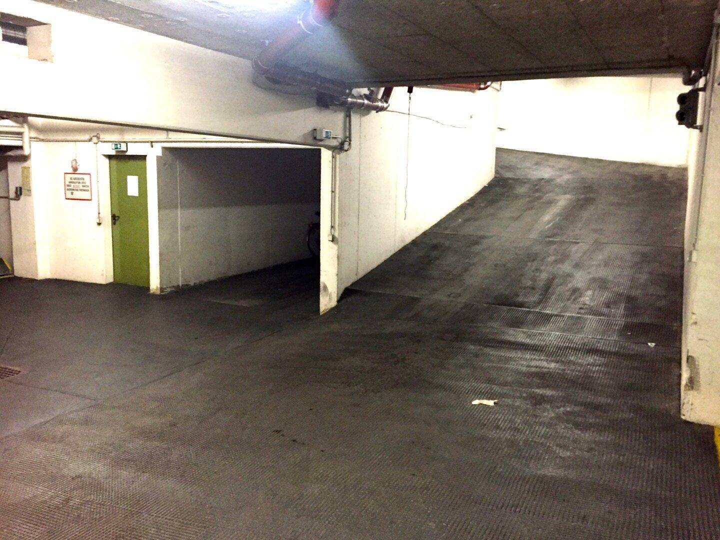 Einfahrt zu einer Tiefgarage mit Rampe und einer grünen Tür zu einem Nebenraum.