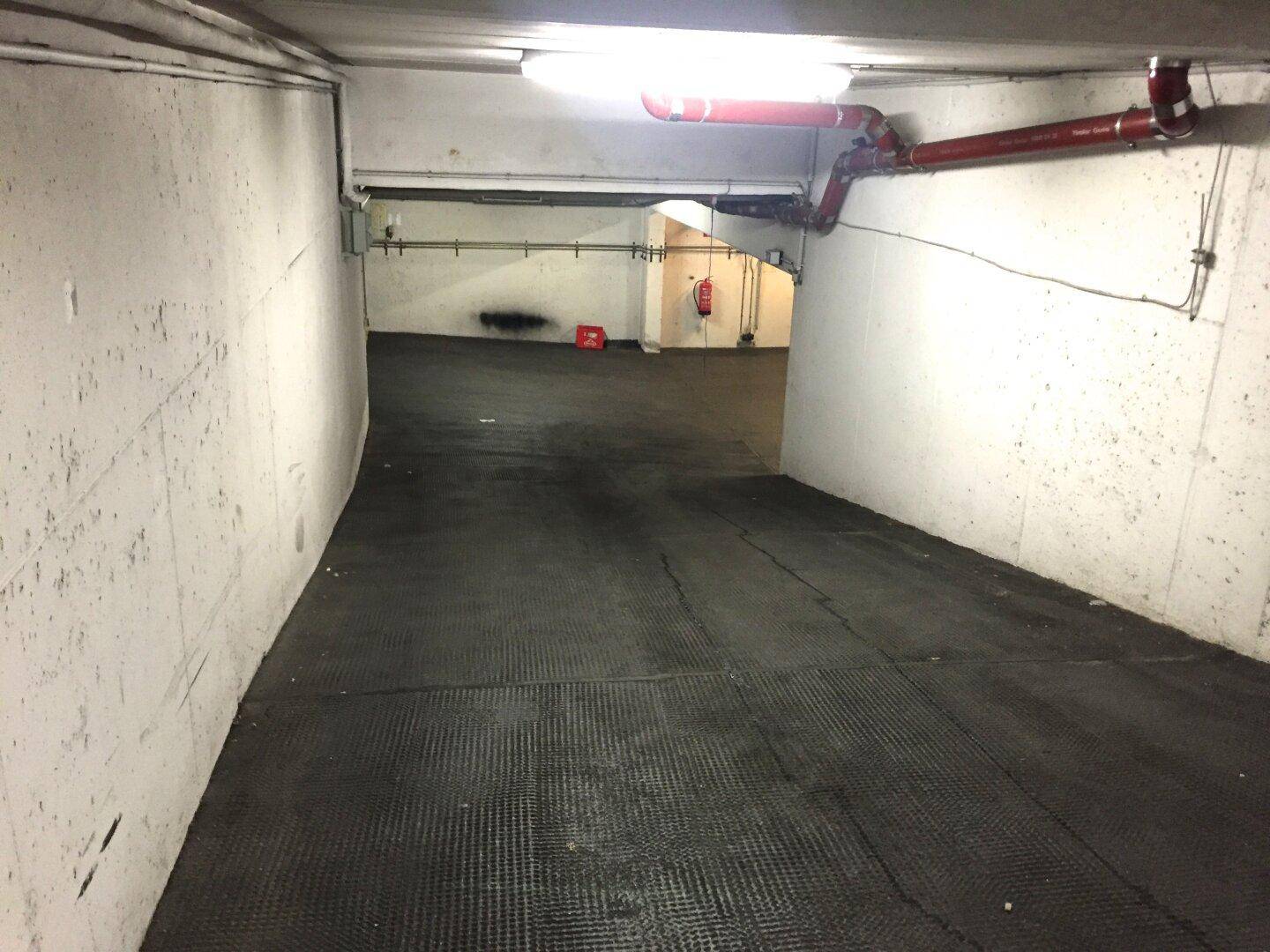 Gang oder Rampe in einer Tiefgarage mit sichtbaren Rohren und einem Feuerlöscher im Hintergrund.