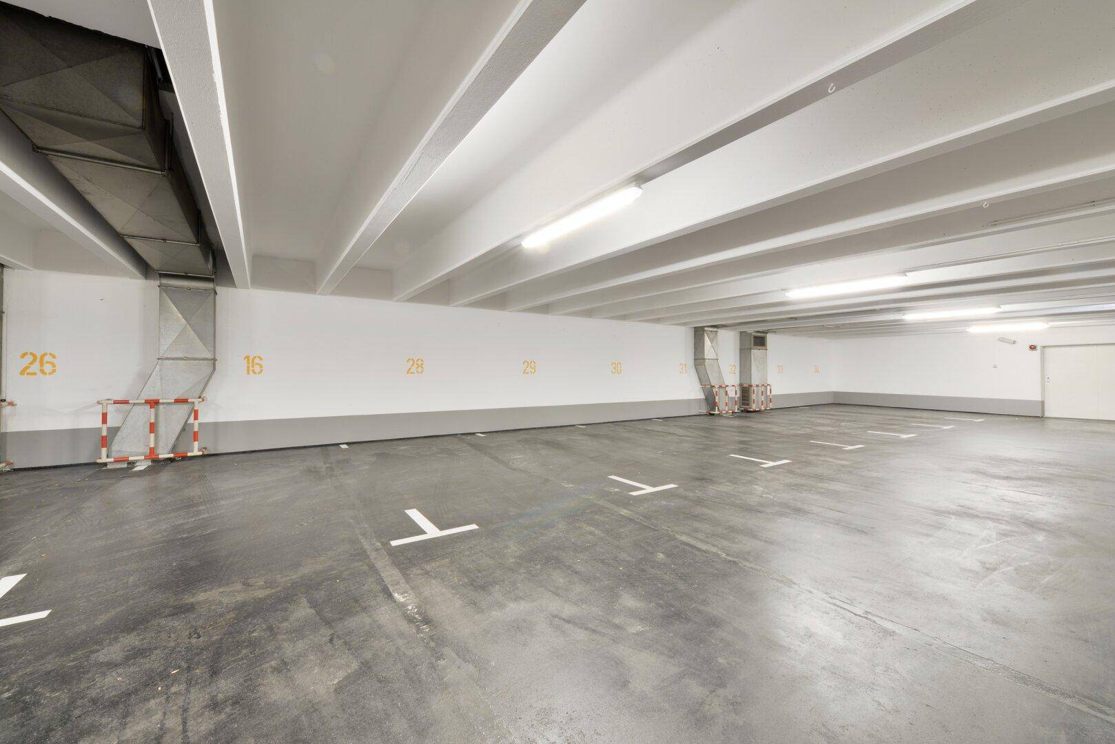 Helle Tiefgarage mit nummerierten Stellplätzen, Betonboden und sichtbaren Lüftungsrohren.