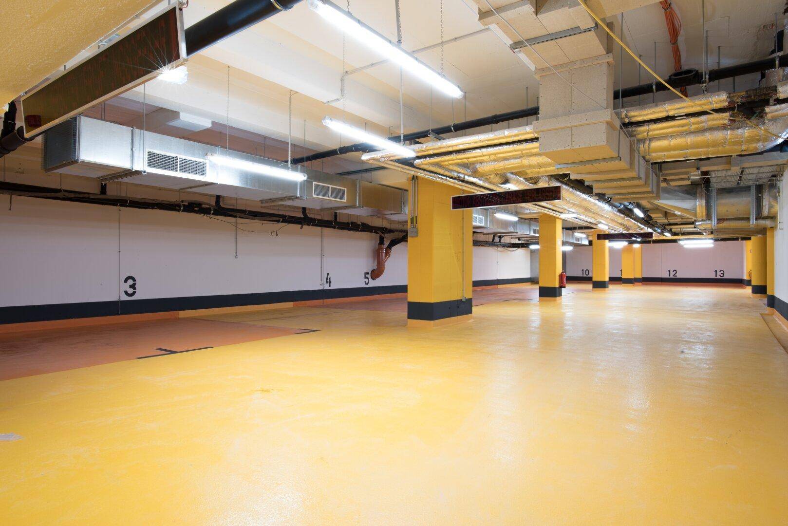 Helle Tiefgarage mit gelbem Boden, nummerierten Parkplätzen und industrieller Deckeninstallation.